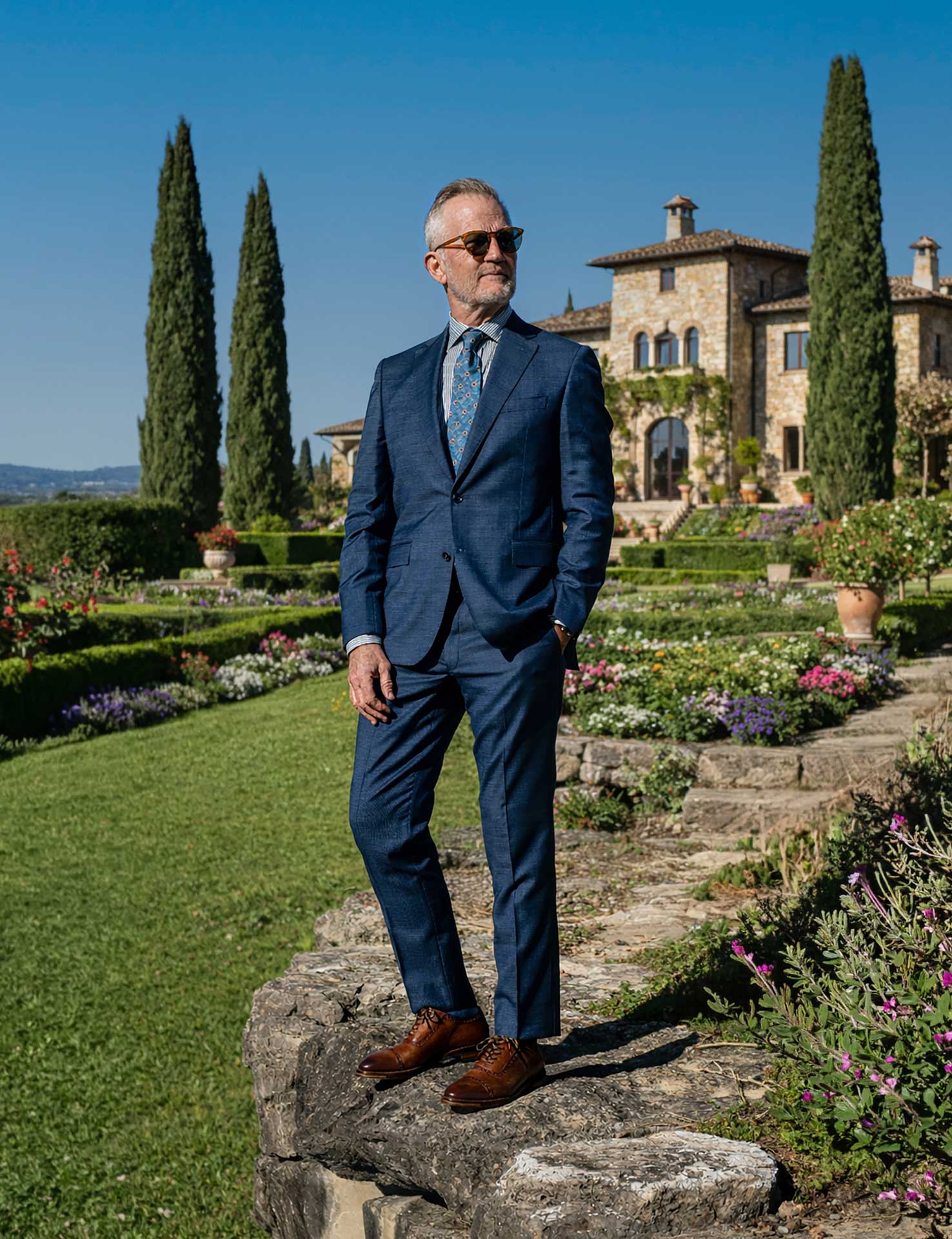 Man wearing a navy suit and brown leather dress shoes on a stone path in a villa garden.