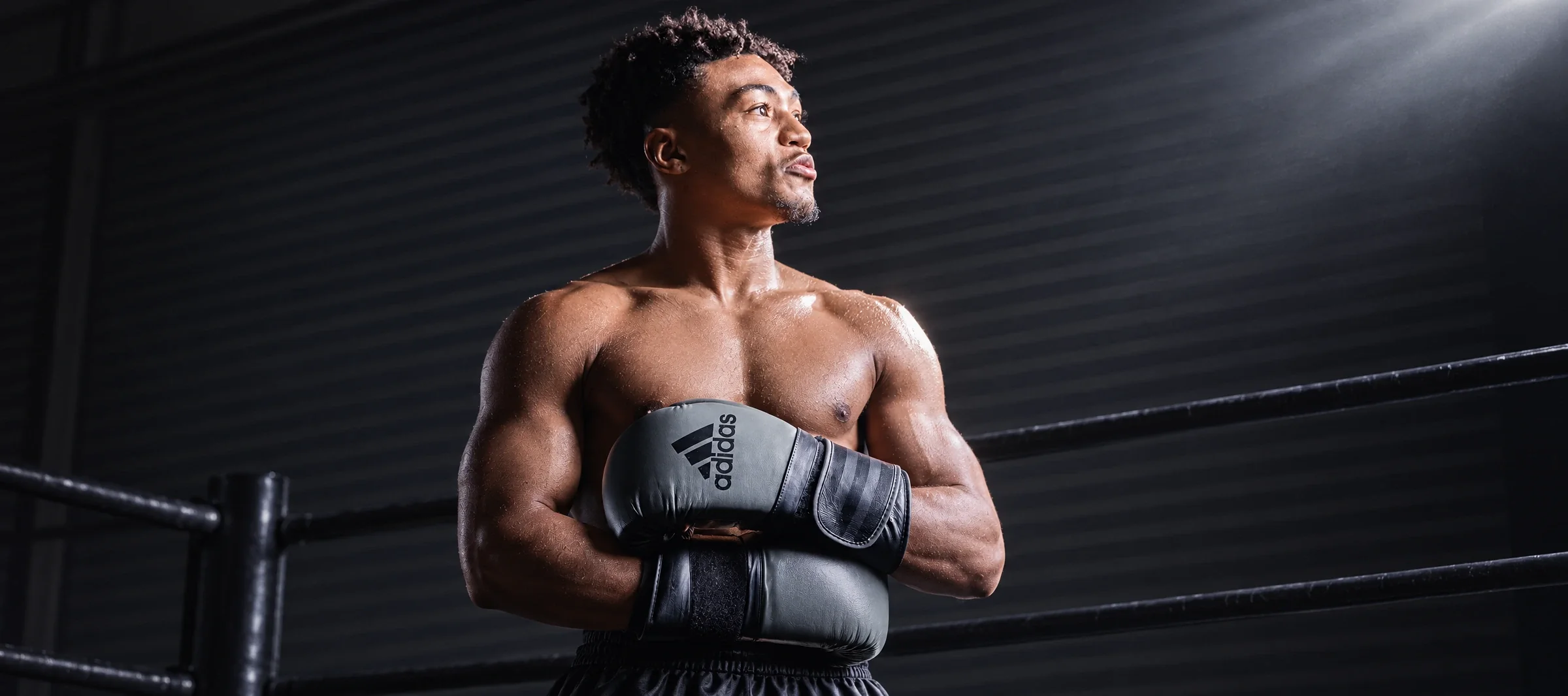 A boxer standing in a dark gym with his arms folded, wearing grey adidas boxing gloves