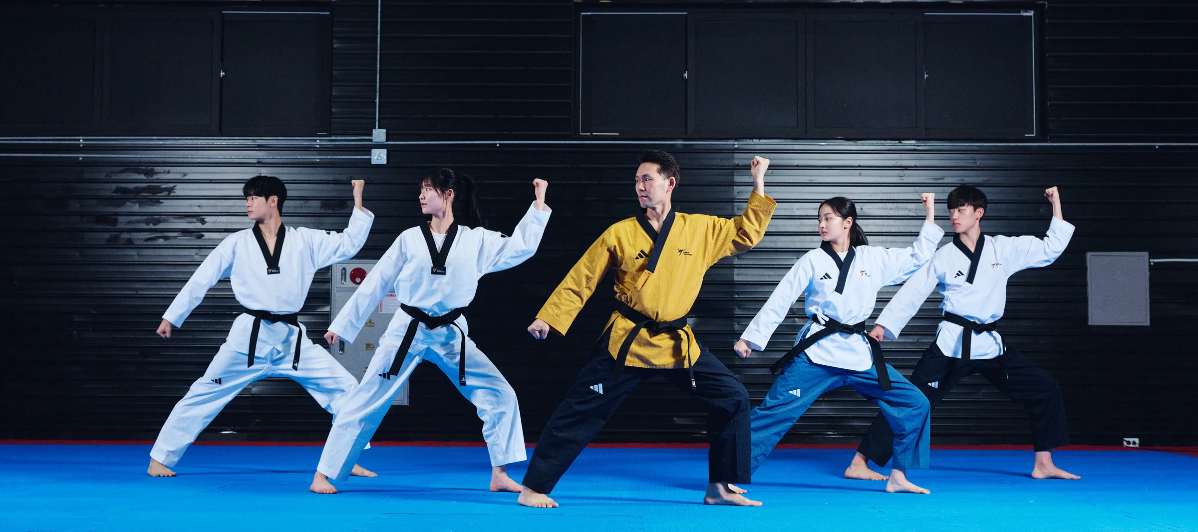 Five taekwondo athletes in adidas poomsae uniforms holding a stance on a blue mat