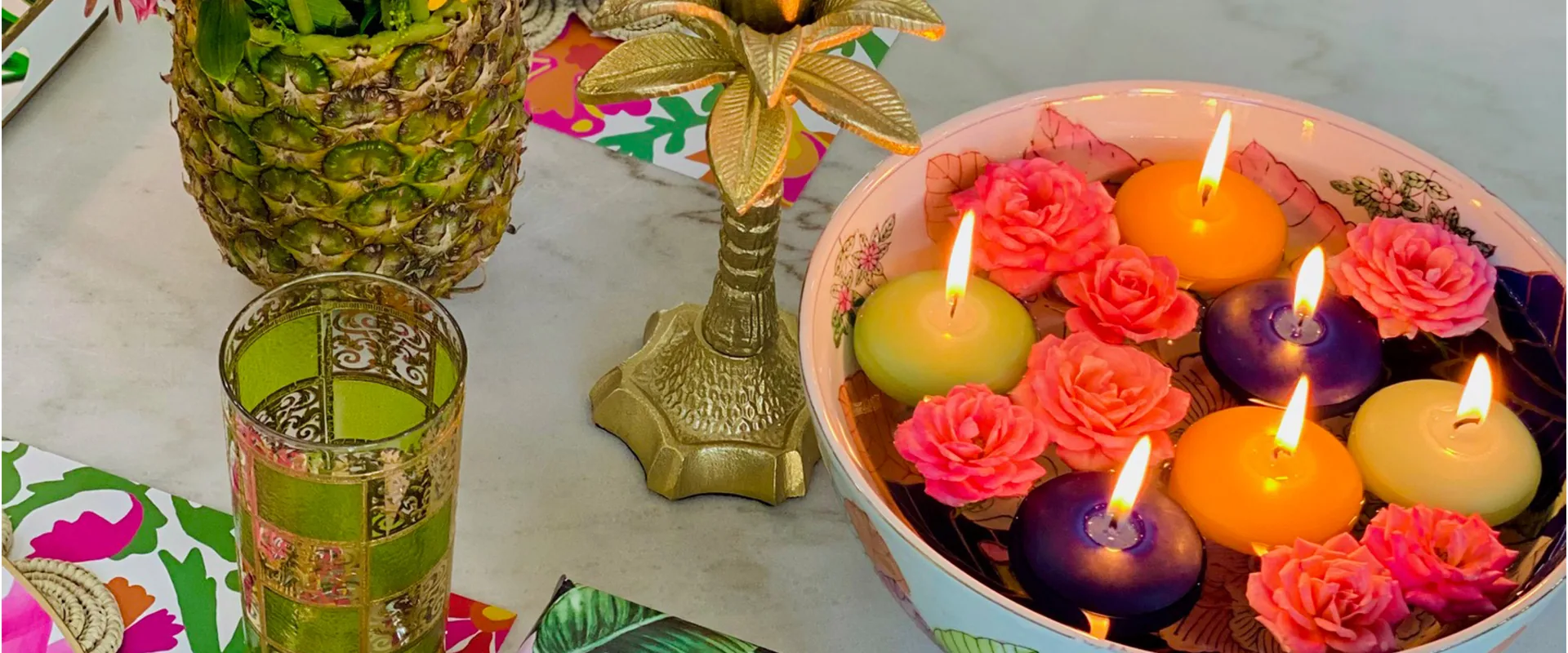 A Creative Candles table setting on marble: floating rose and pillar candles lit in a footed bowl, beside a gold palm candlestick and a carved green votive