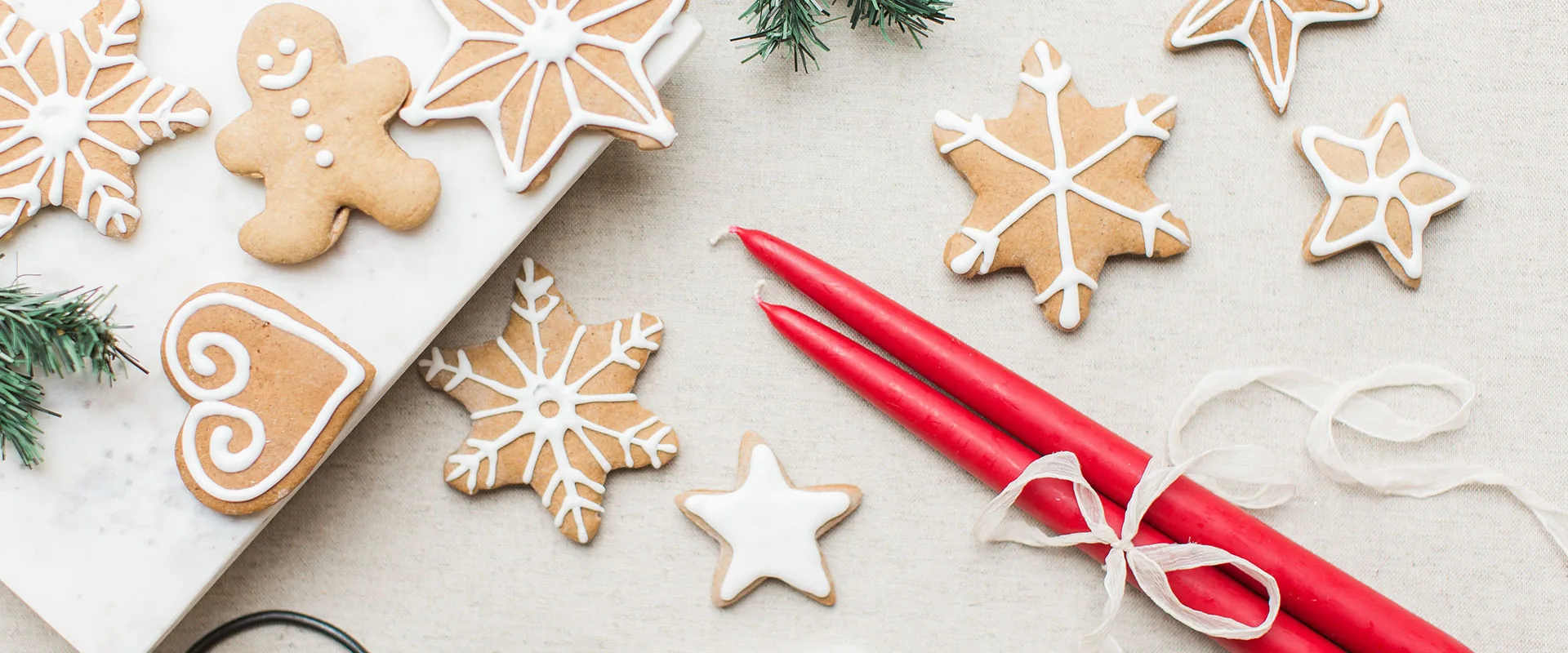 Iced gingerbread snowflakes and stars on a marble board beside two red taper candles tied with ribbon, pine sprigs on linen