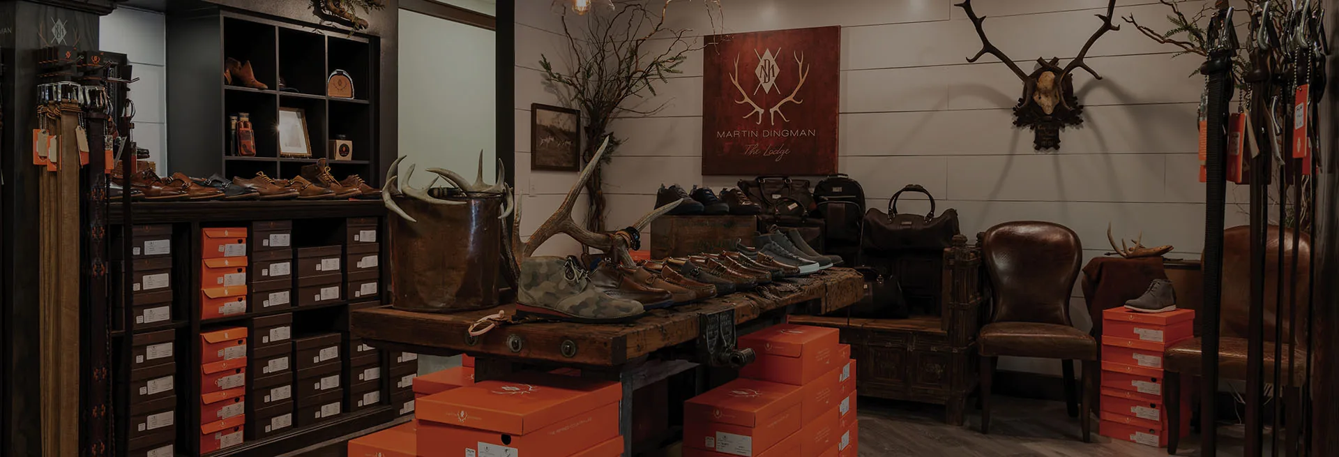 The Martin Dingman lodge shop, shoes and boots on a reclaimed timber table under the antlered wordmark, orange boxes stacked below