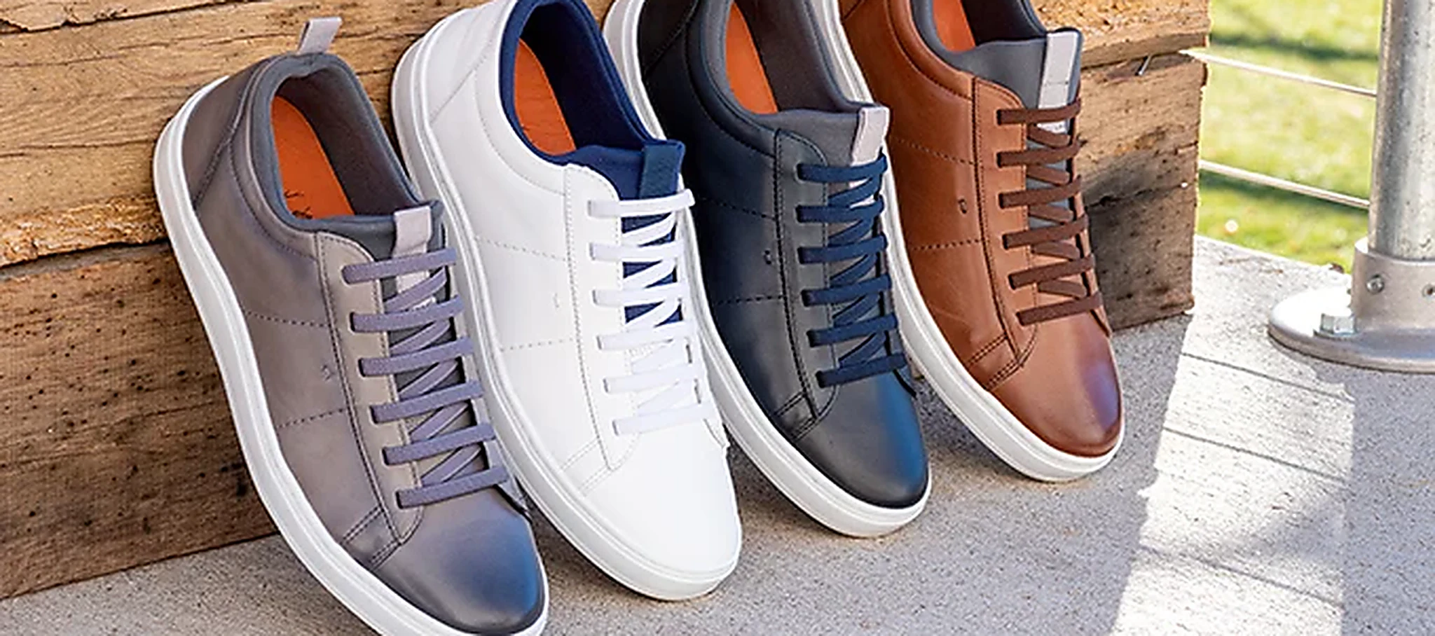 Four Martin Dingman leather sneakers in grey, white, navy and cognac, lined up against a weathered timber wall in sunlight