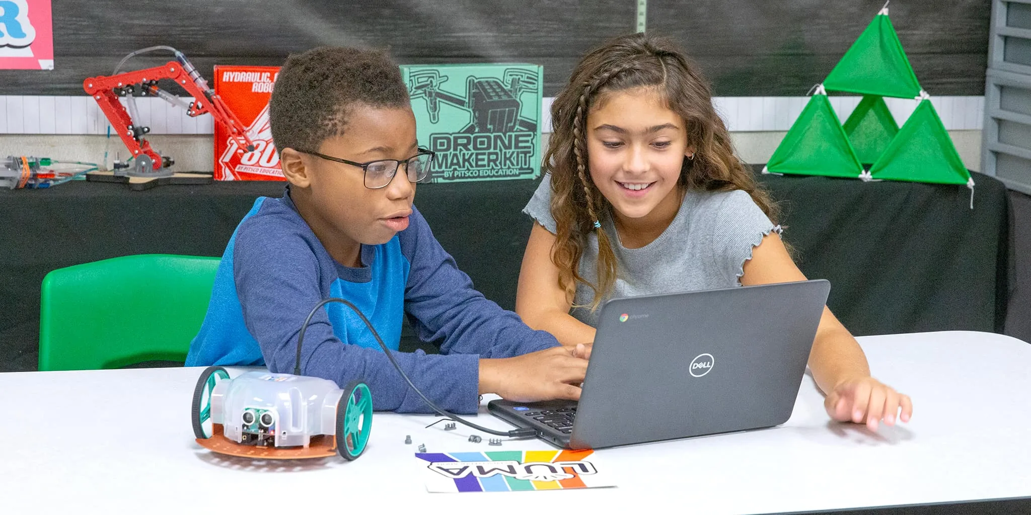 Two students at a classroom table, one typing on a laptop while the other looks on, a wheeled Pitsco robot and a UUMA card in front of them and Drone Maker Kit and hydraulic arm builds on the shelf behind