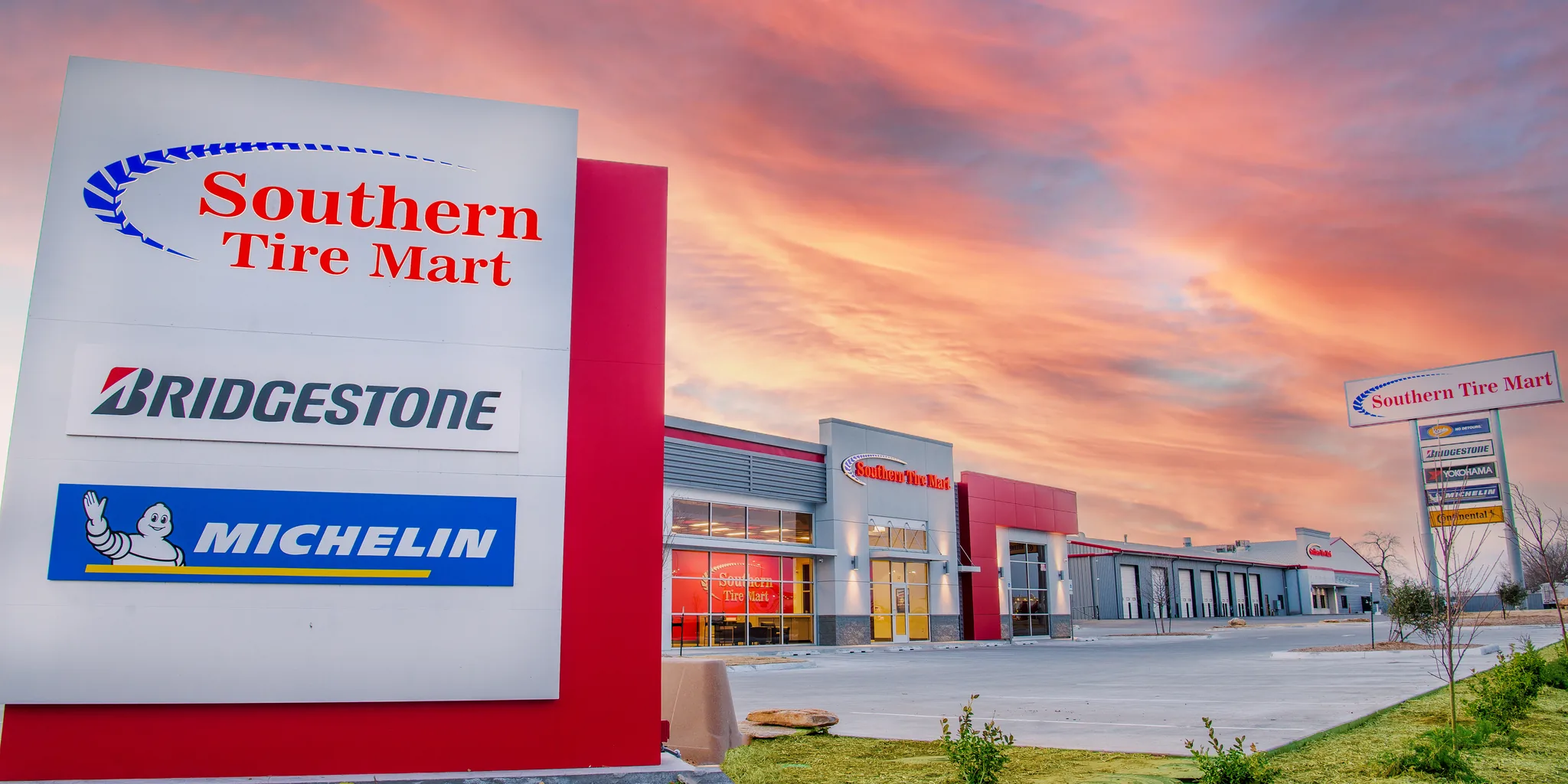 A Southern Tire Mart store at sunset, its pylon sign carrying the Southern Tire Mart, Bridgestone and Michelin names in the foreground and the lit showroom and service bays beyond under a pink and orange sky