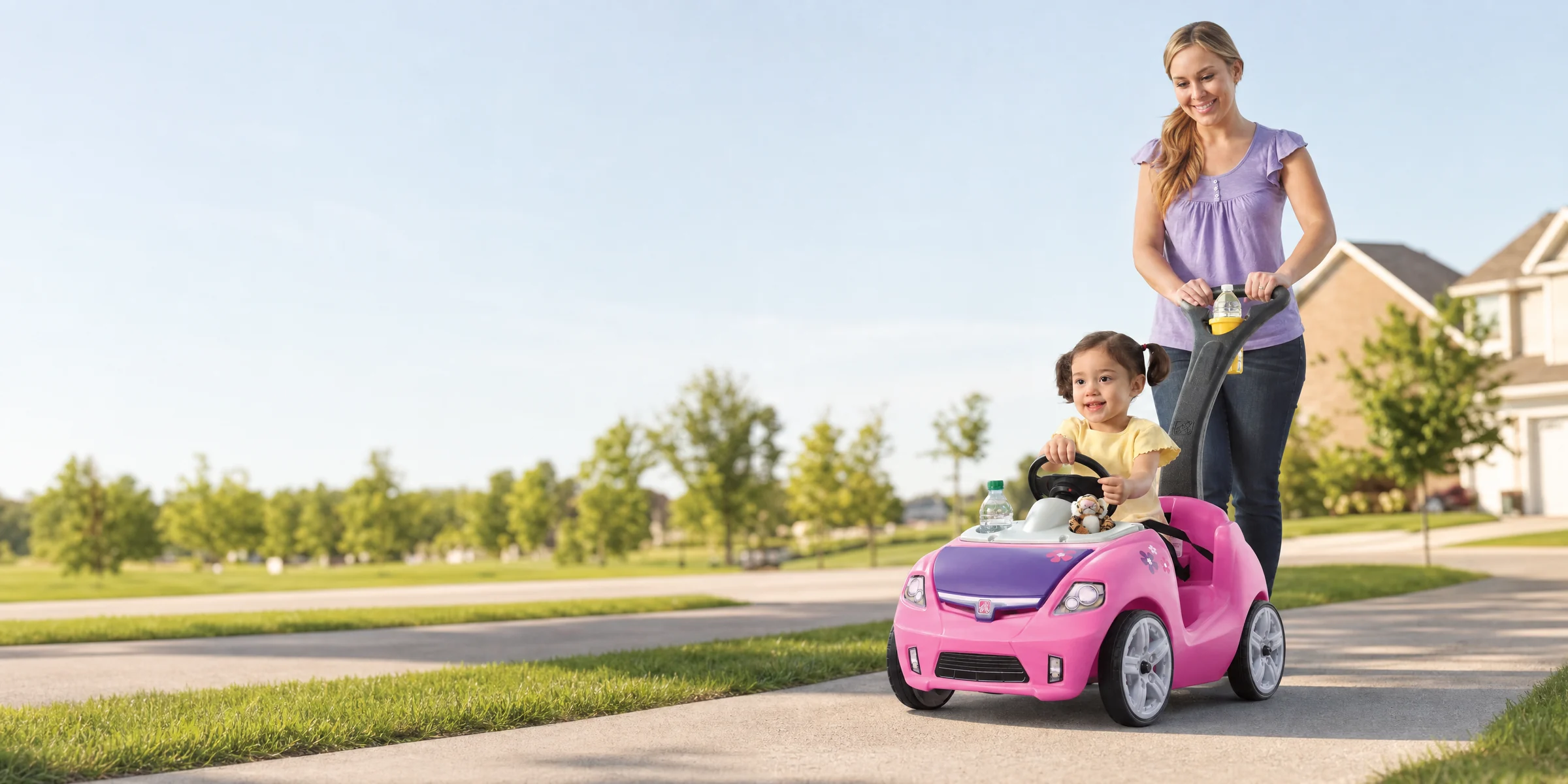 A mother pushes a smiling toddler in a pink Step2 Whisper Ride II push car along a neighborhood sidewalk