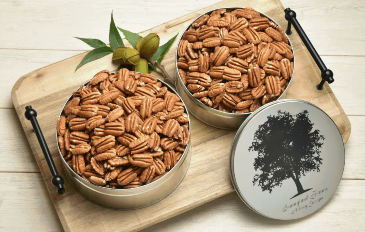 Two Sunnyland Farms gift tins filled with pecan halves on a wooden board, beside a tin lid stamped with the Albany, Georgia tree mark